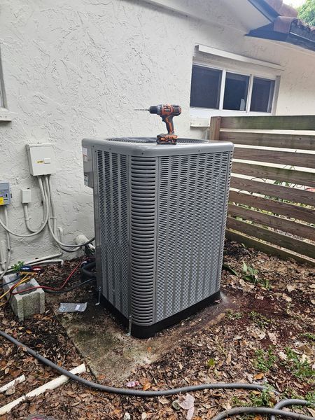 An air conditioning unit with a power drill on top, outside a building, surrounded by foliage.