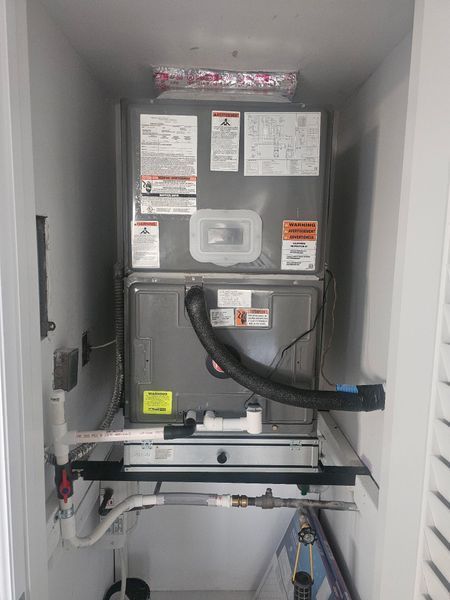 HVAC unit in a closet. Gray metal box with labels and black vent hose connected. White pipes and a black frame are below.