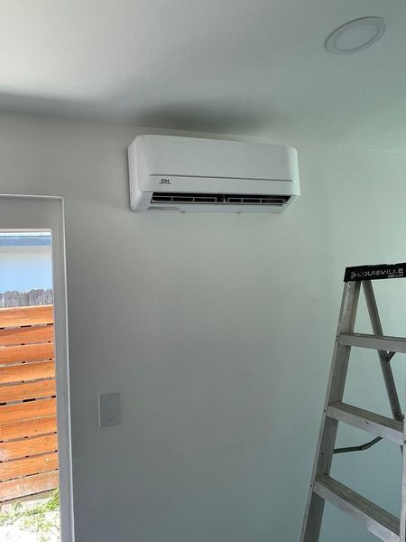 White wall-mounted air conditioner. A ladder stands nearby; a doorway and ceiling light are also visible.