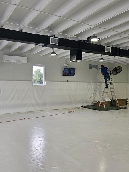 Man on ladder works on a unit in a large, white-walled indoor space. Air vents, lights, and a window are visible.