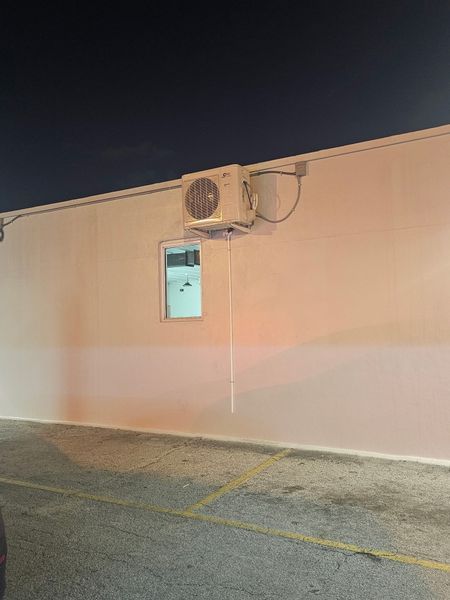 Air conditioner unit mounted on a white building wall with a small window below, set against a dark night sky.