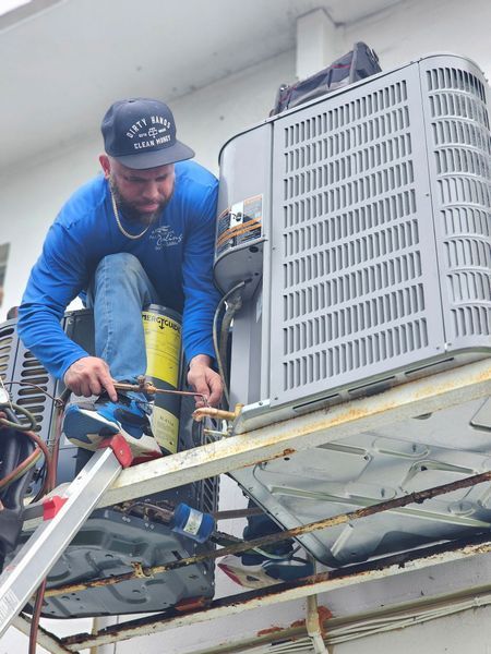 HVAC technician in blue shirt and hat working on an air conditioning unit outside, using tools.