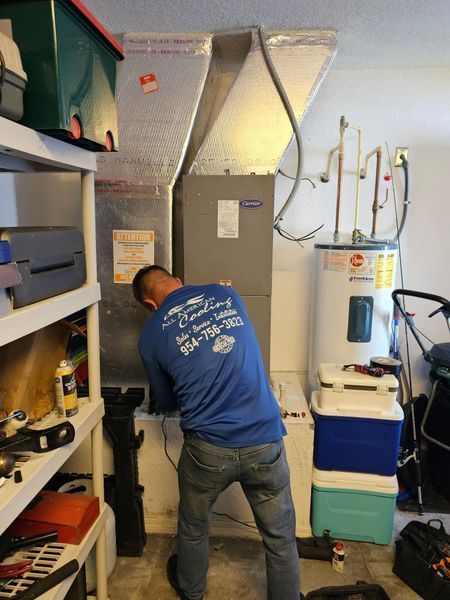 Man in blue shirt working on HVAC unit in a garage, next to a water heater and shelving.