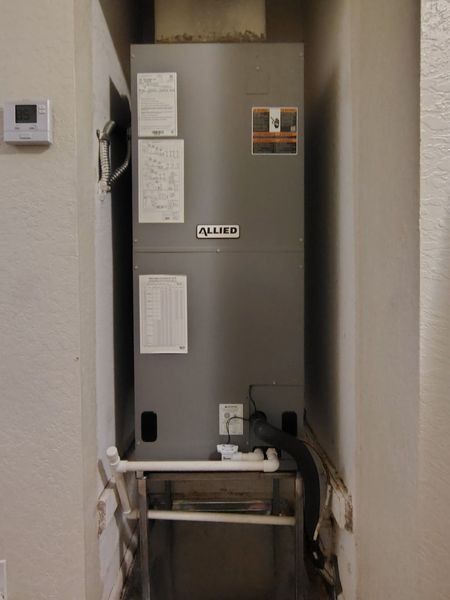 A gray HVAC unit in a wall alcove with attached white pipes, a thermostat is visible on the wall.