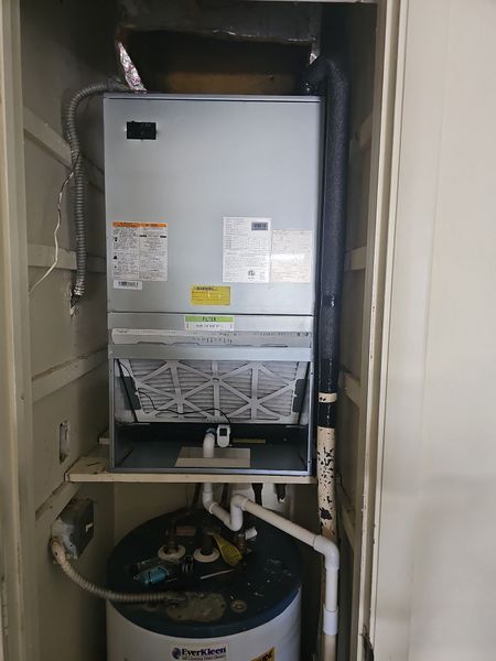 Furnace and water heater inside a utility closet. Silver and white appliances. Black flue pipe. White and gray walls.