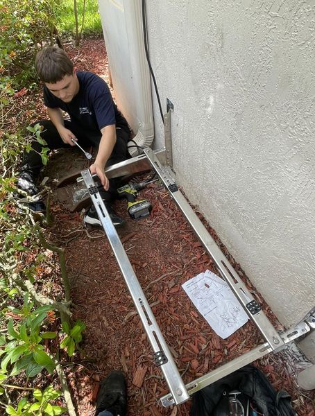 Person kneeling, installing an AC unit bracket on a building's exterior wall. A drill and parts are visible.