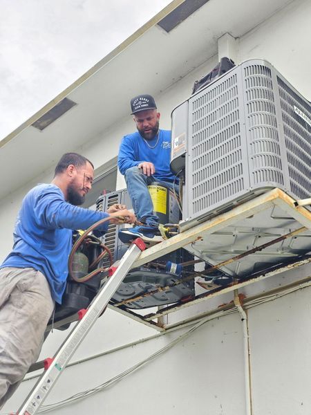 Two HVAC technicians on a rooftop platform, working on an air conditioning unit.