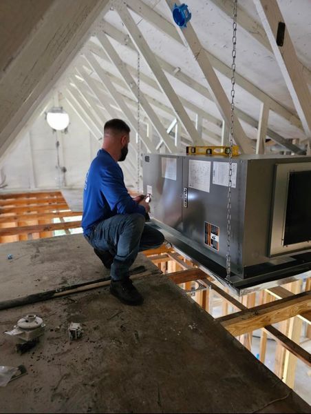 Man squatting, inspecting HVAC unit suspended in attic, using a level.