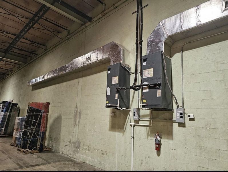 Two HVAC units mounted on a concrete wall in an industrial setting, with ductwork and electrical components visible.