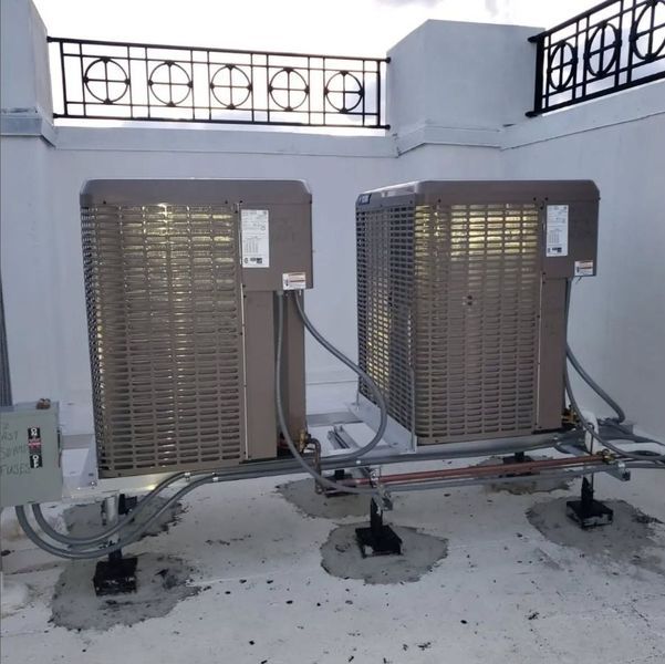 Two rooftop air conditioning units. Grey metal, atop concrete pads, next to a black railing.