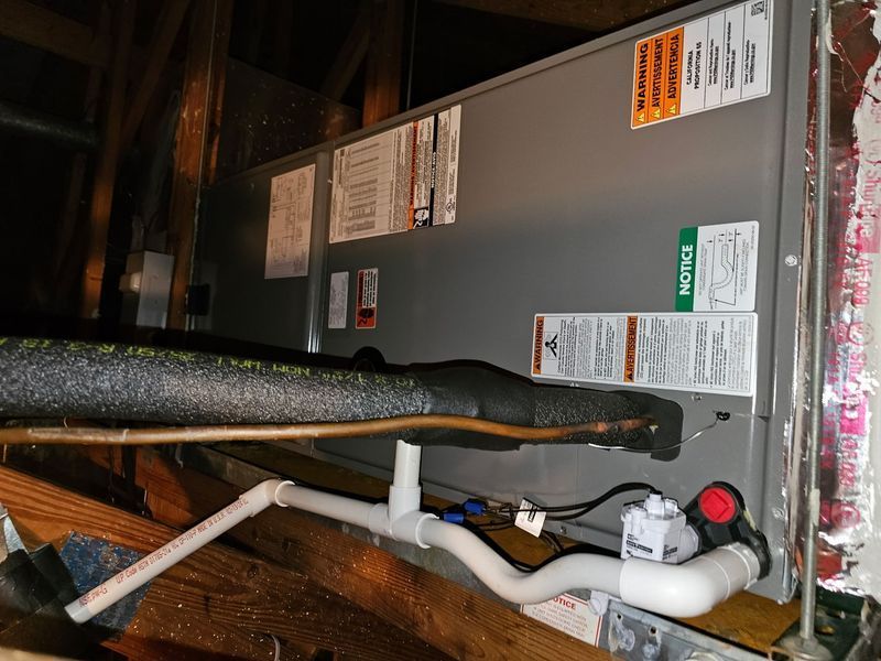 An HVAC unit in an attic with insulated pipes, and condensate pump, and warning labels.