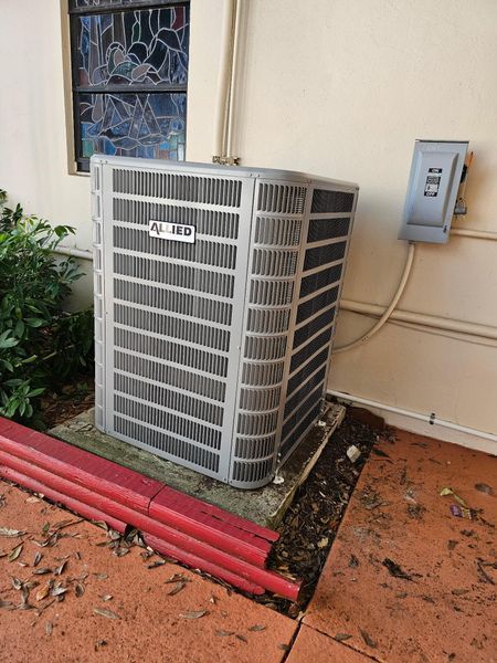 An air conditioning unit next to a building wall with a stained glass window and electrical box.