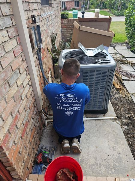 HVAC technician kneeling by an air conditioning unit next to a brick wall, installing or repairing it.