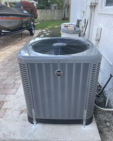 Gray air conditioning unit on a concrete patio next to a house, with a boat in the background.
