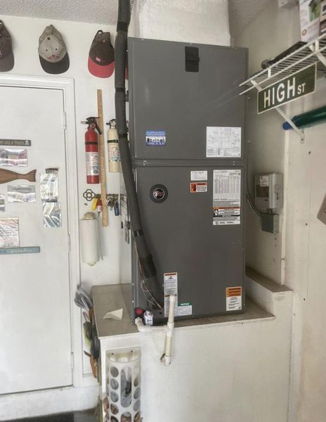 HVAC unit installed inside a garage, next to a door, fire extinguisher, and various items. Gray unit.