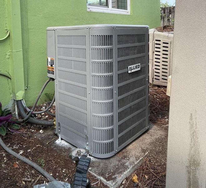 Gray air conditioning unit on a concrete pad near a green wall and a window.