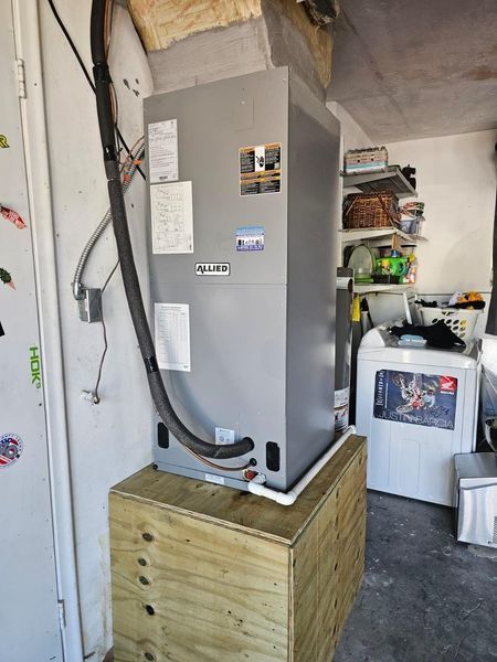 HVAC unit on a wooden platform in a garage. Grey unit, black tubing, white door, and a washing machine are visible.