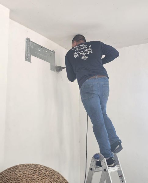 Man on a ladder drills into a wall to install an air conditioner bracket; indoor setting.