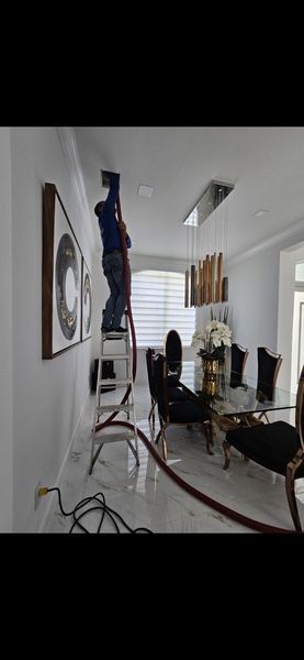 Person on ladder working on ceiling in a dining room. Red hose and electrical cord on floor.