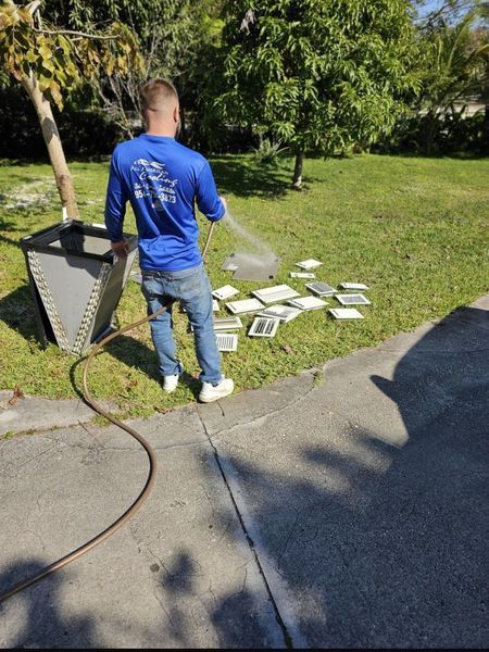 A person in blue shirt and jeans sprays water on tiles on a grassy area, near a driveway and a large metal object.