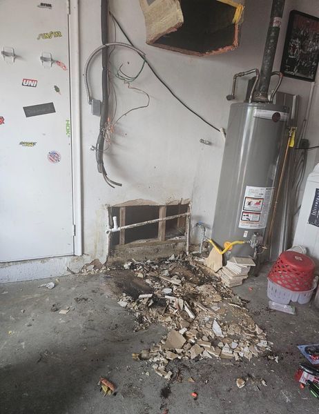 Damage to garage wall with exposed framing and debris. Water heater nearby.