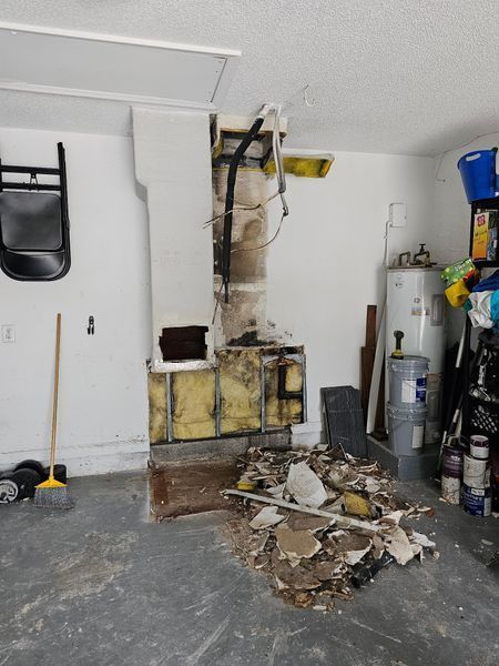 Garage with exposed chimney and debris on the floor, next to a water heater and cluttered shelves.