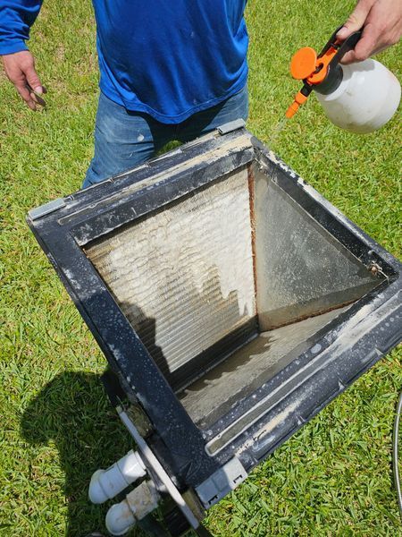 Person sprays cleaning solution on an air handler filter box, outdoors on grass.