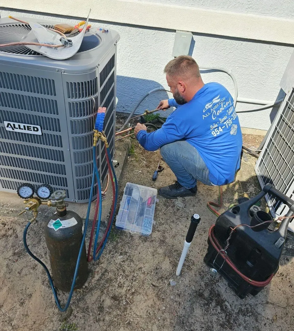 HVAC technician servicing an Allied air conditioning unit outdoors, using gauges and tools.