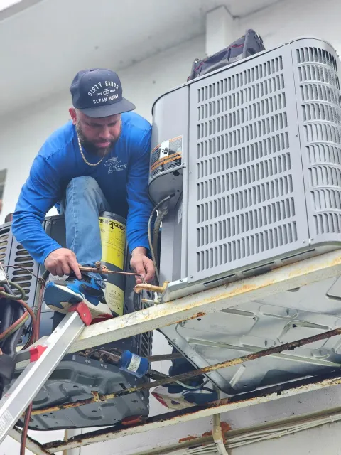 HVAC technician working on an air conditioning unit outside. He wears a hat, long sleeve shirt, and jeans while soldering.
