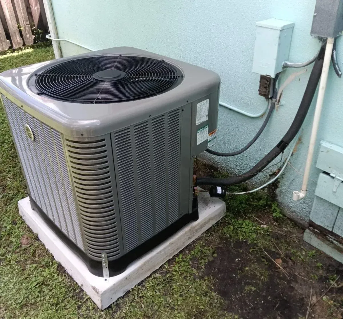 Outdoor air conditioning unit, gray metal, on concrete pad, next to blue house wall and electrical box.