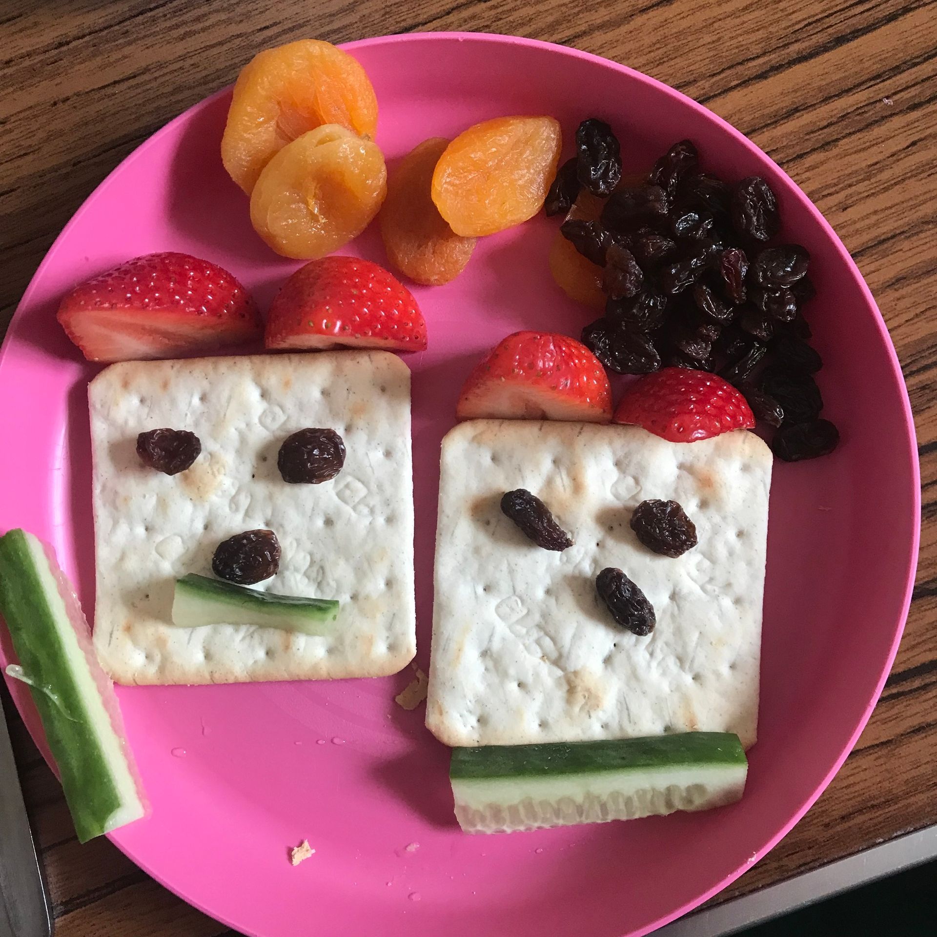 A pink plate with crackers raisins and strawberries on it