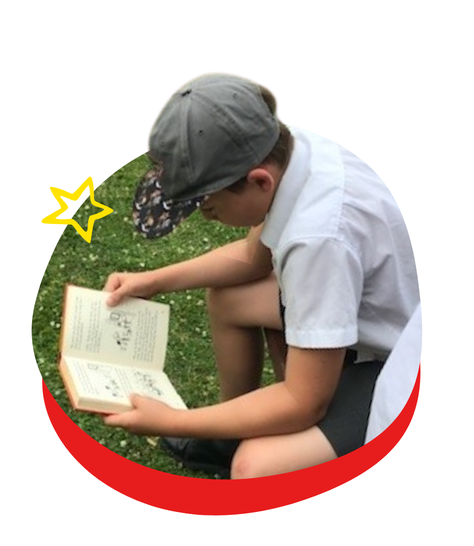 A boy wearing a hat is reading a book in the grass