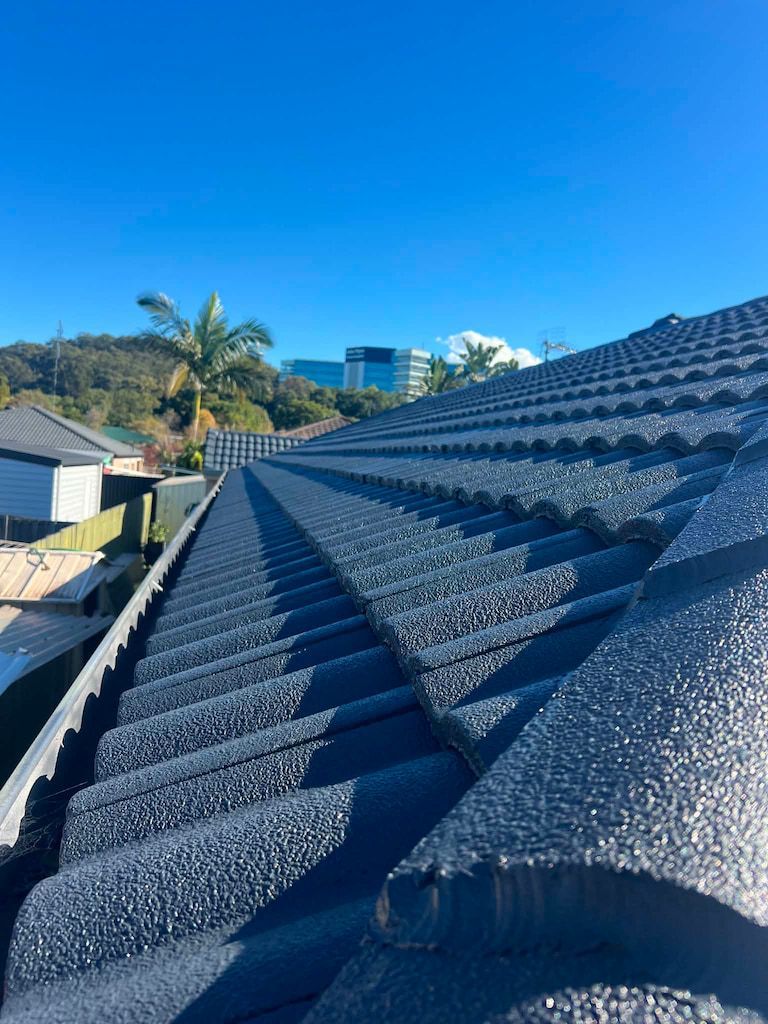 Close-up of a dark gray tiled roof under a bright blue sky with palm trees in the background