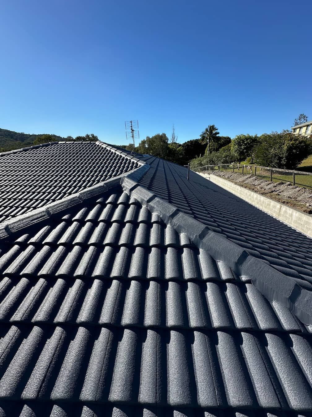 Roof with dark gray tiles under a clear blue sky, viewed from a rooftop ridge.