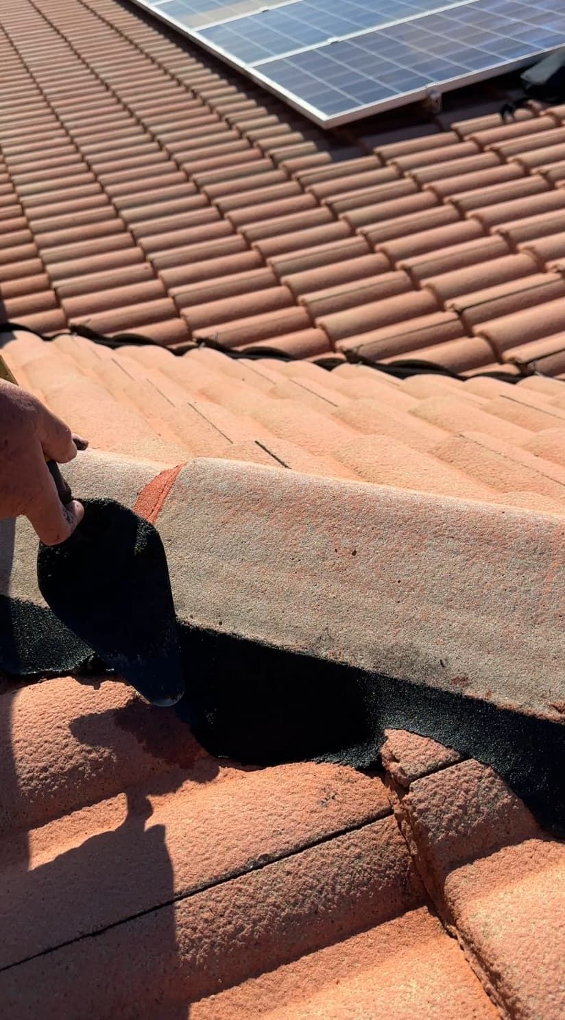 A person uses a trowel to apply black sealant to a gap in terracotta roof tiles near solar panels.