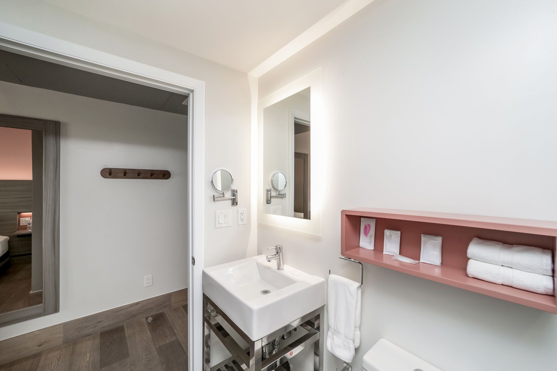 Modern bathroom with a white sink, mirror, and pink shelving.  Doorway to a bedroom is visible.