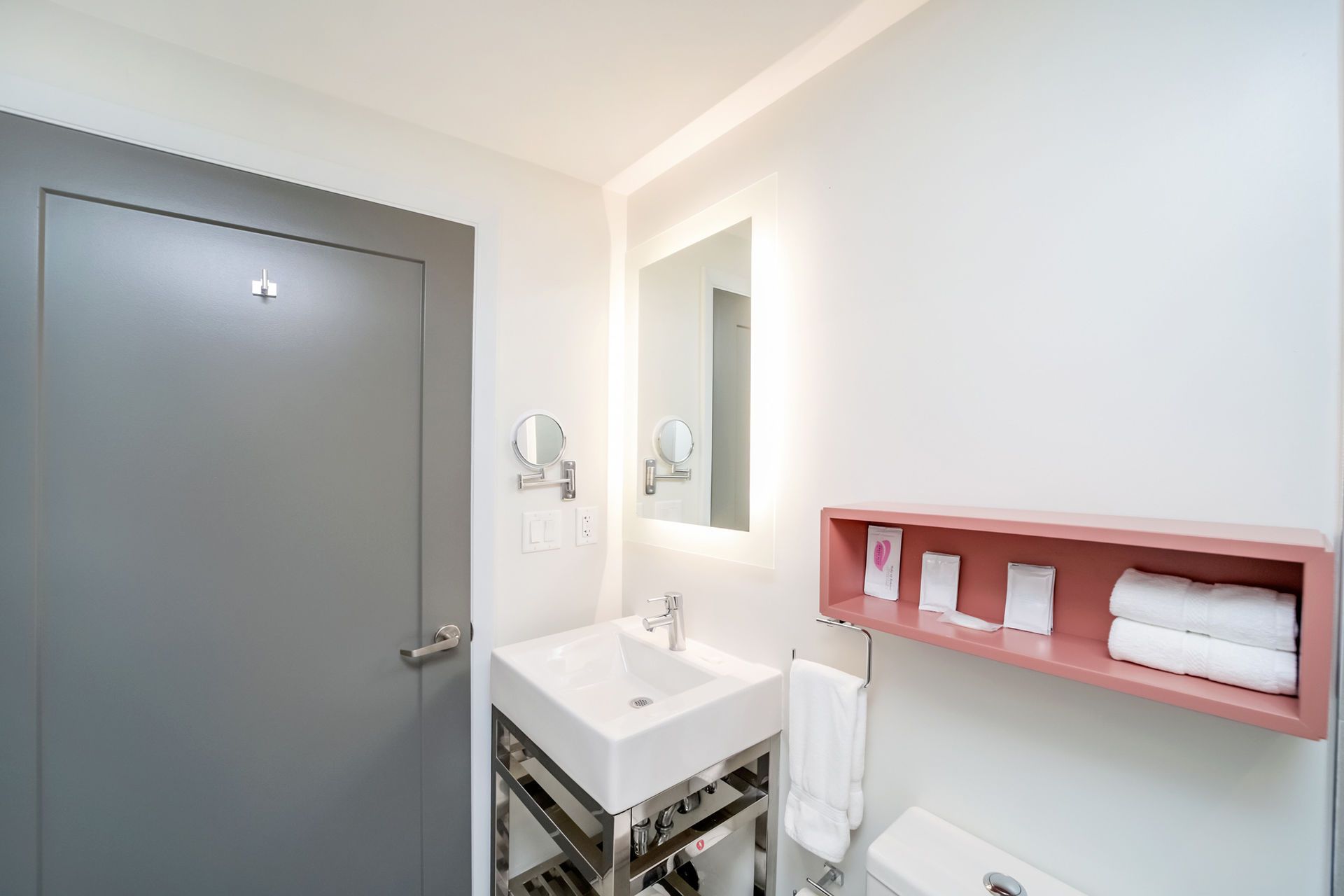 Bathroom with white walls, gray door, square sink, pink shelf with towels, and illuminated mirror.