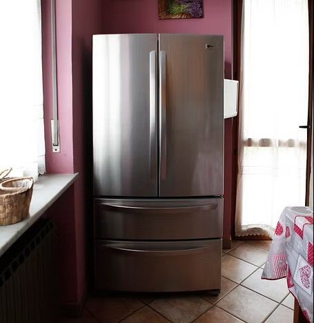 Frigorifero in acciaio inox con porta alla francese in una cucina con pareti rosa, accanto a una finestra e a un tavolo.