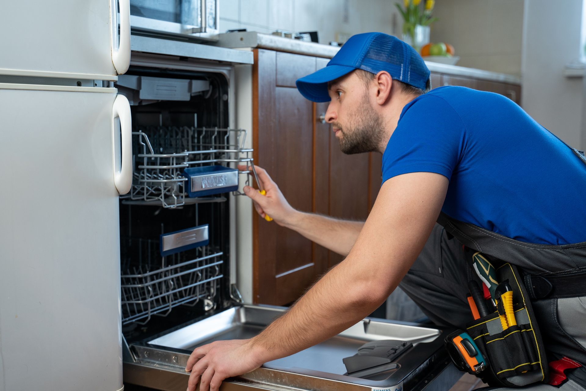 A worker in a blue uniform performing an appliance unit repair, repairing a dishwasher in a kitchen.