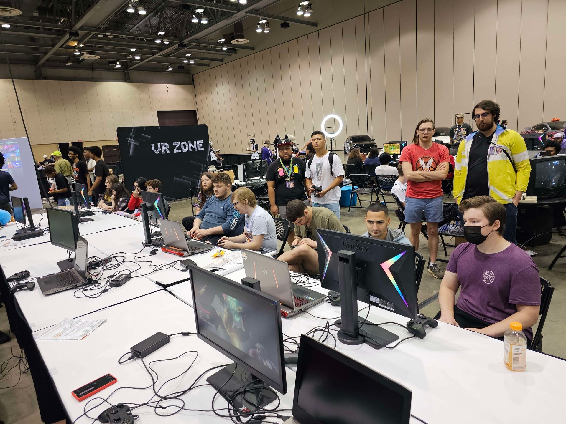 People at gaming event, seated at tables with computers and monitors; some watching, one in mask. 