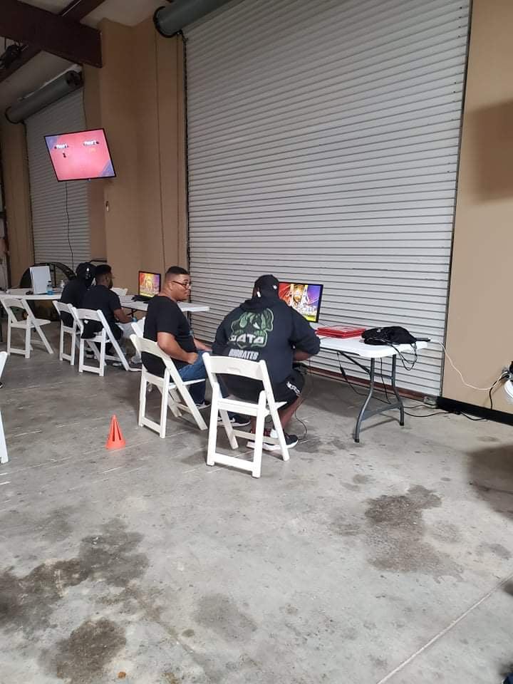 Three people seated at tables, watching a screen in a garage-like space; one table has a small orange cone on it.
