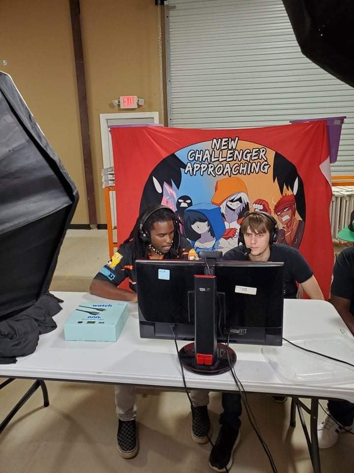 Two people at a table, playing a video game at an event. A banner behind them reads 