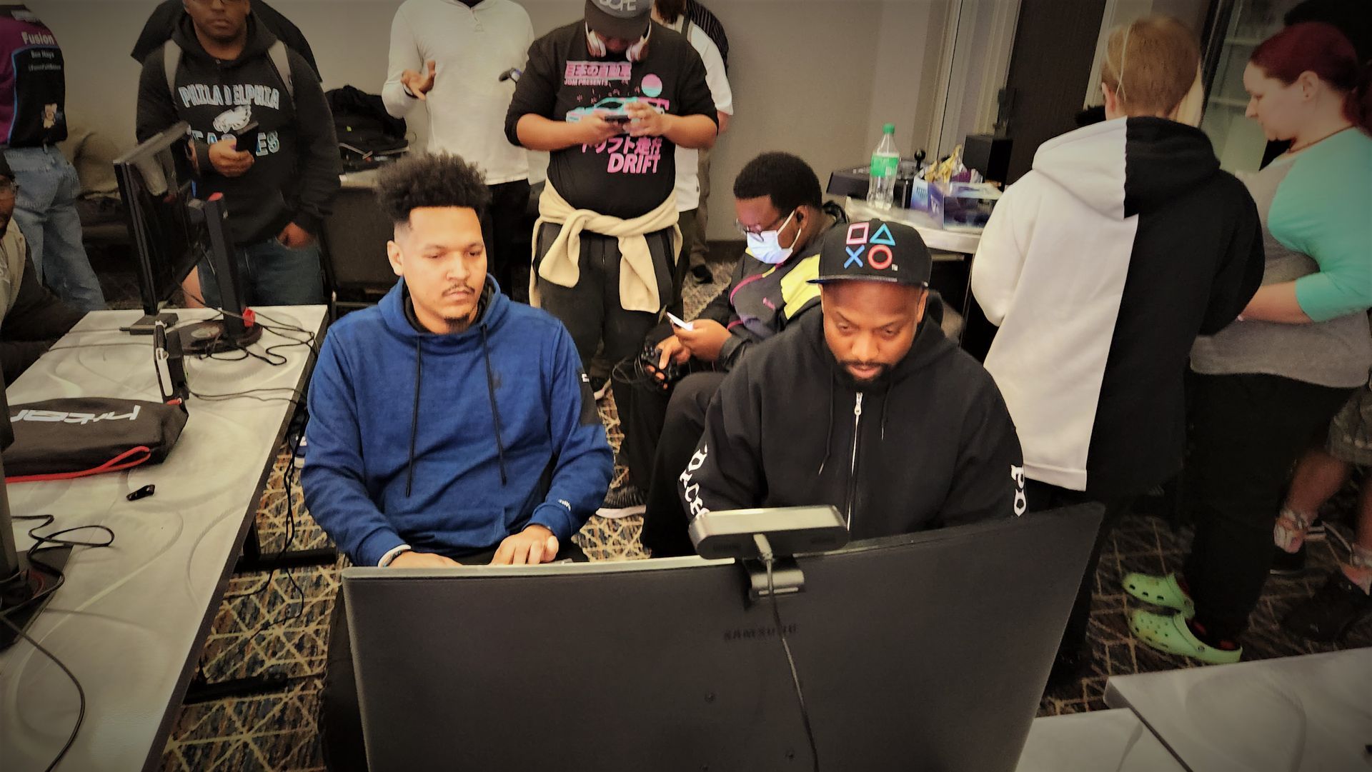 Two men playing video games at a table with monitors, surrounded by other people in a room.
