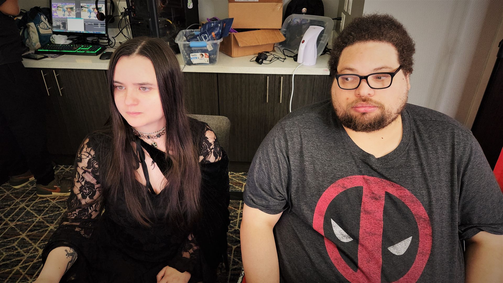 Two people sitting: woman in black lace, man in Deadpool shirt with glasses. Inside room.