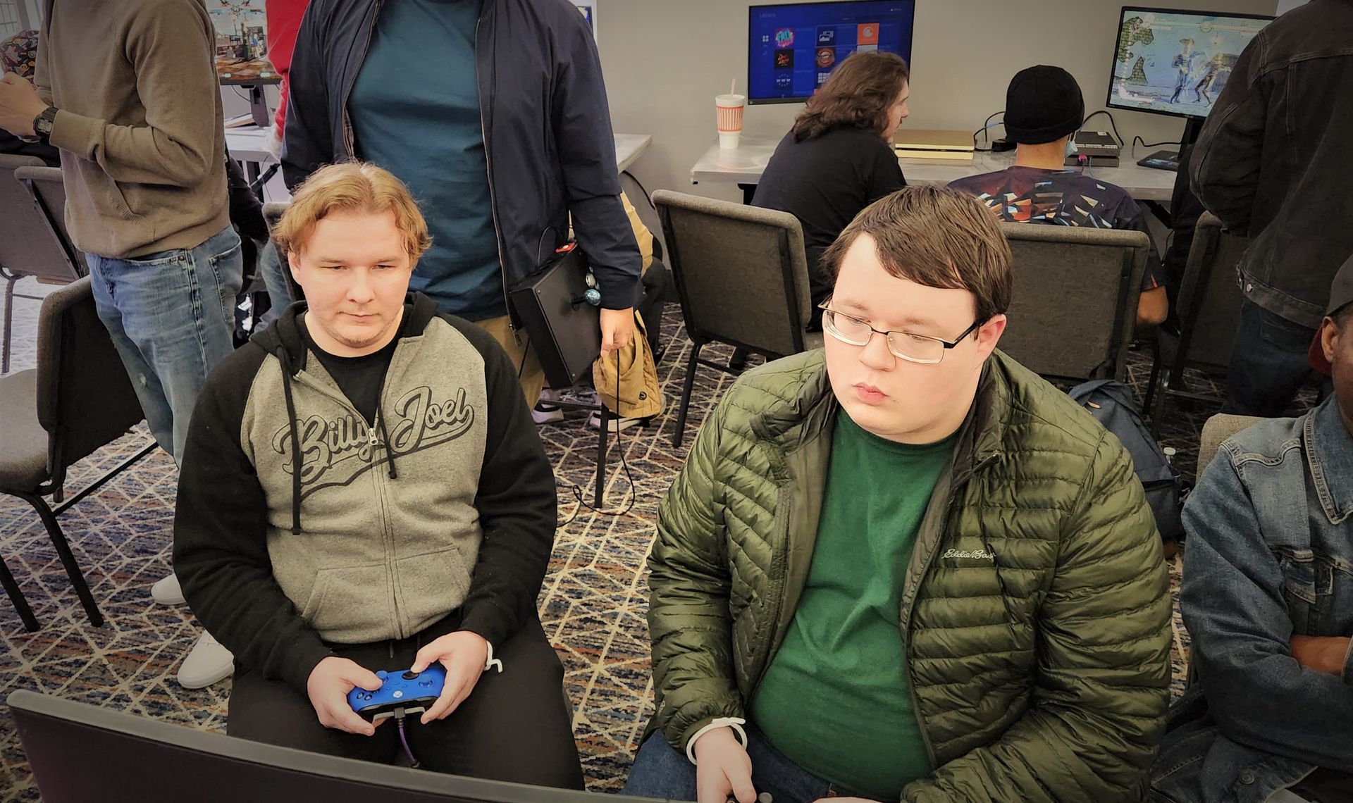 Two men sit playing video games; one holds a controller. Other people are in the background.