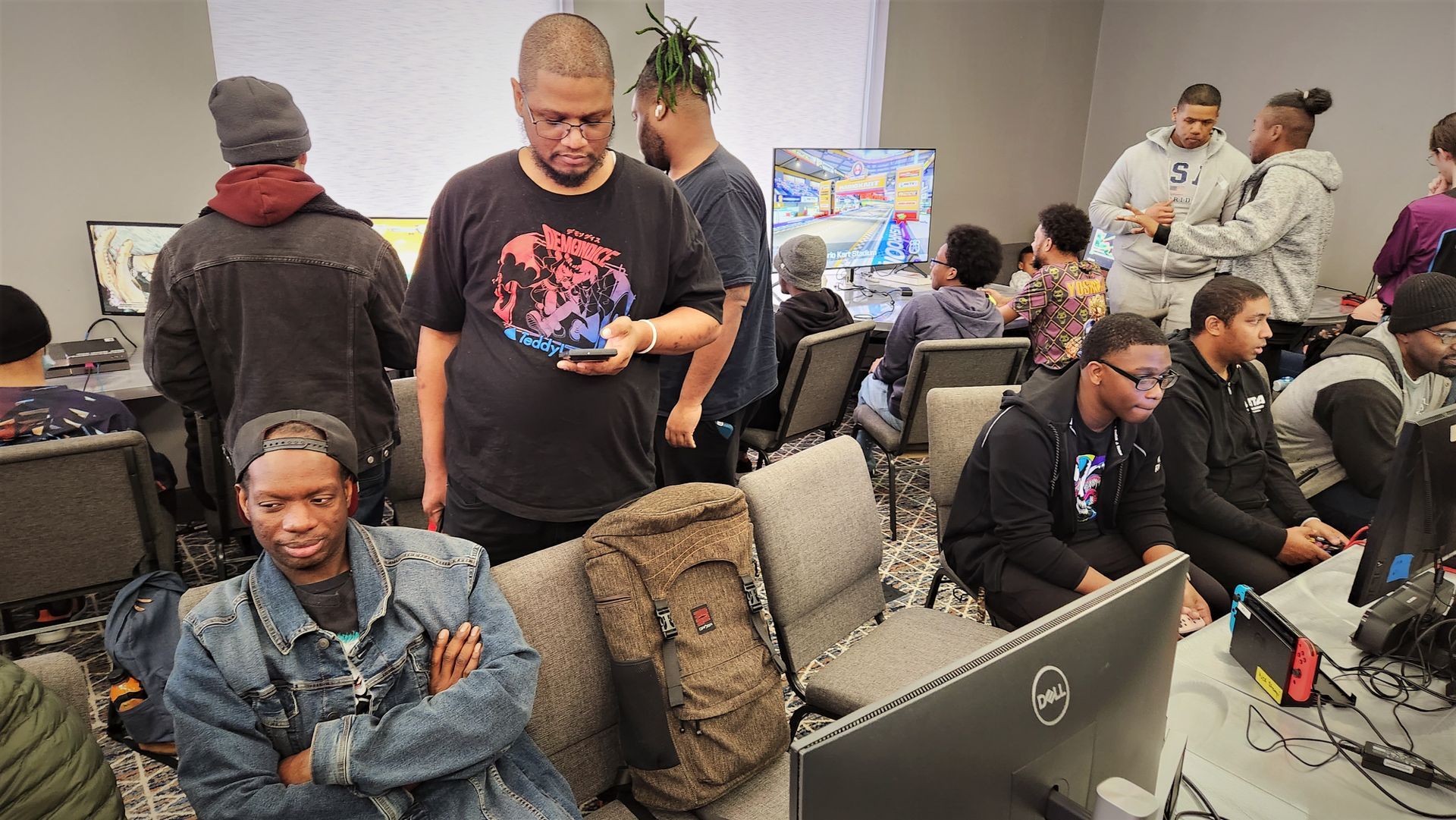 People gaming in a room, some watching screens. Several men are present, some using phones or talking.