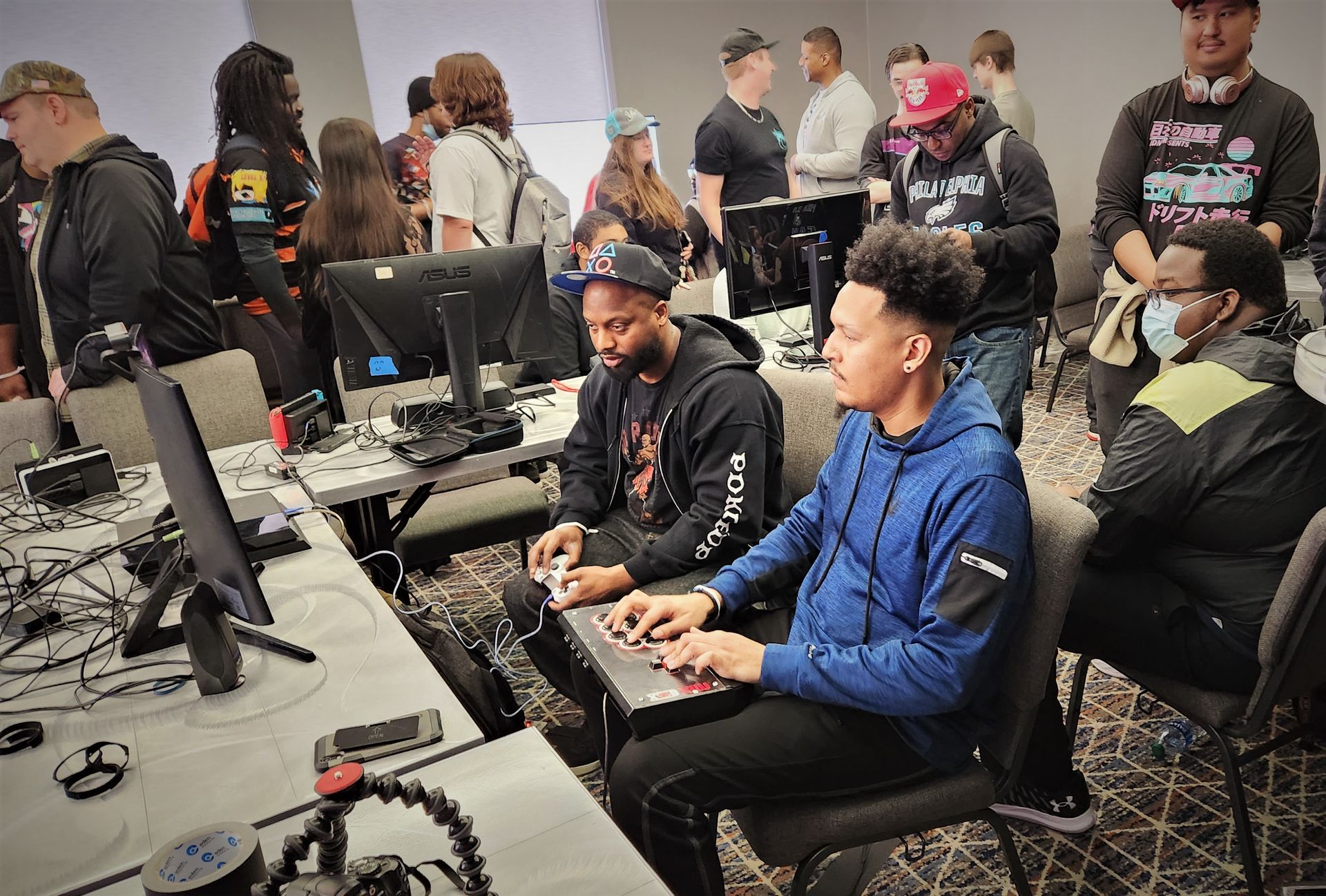 Two men playing video games at a gaming event, using arcade-style controllers, surrounded by other attendees.