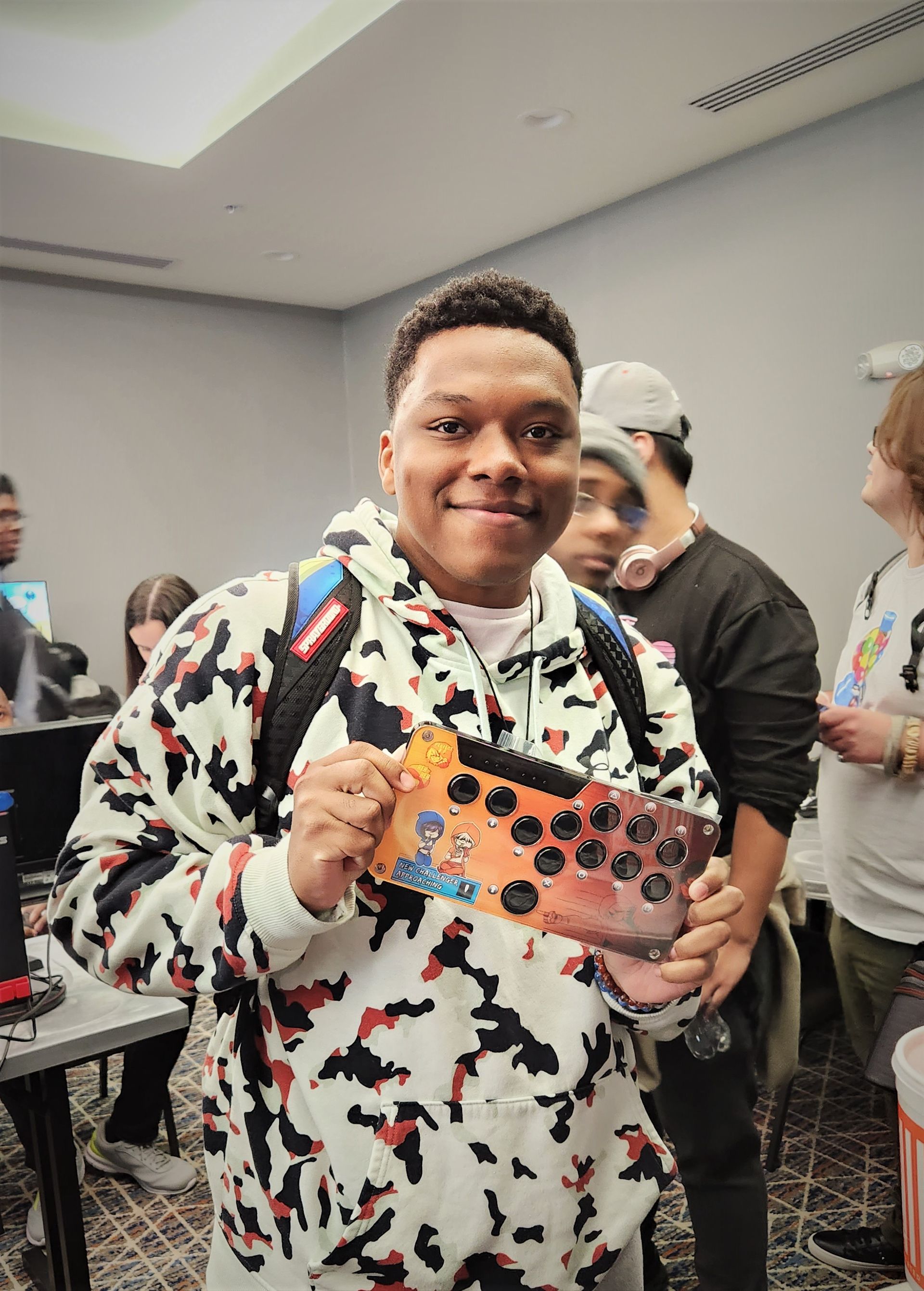 Person holding a customized game controller, smiling. They wear a camo hoodie, and stand in a room with other people.