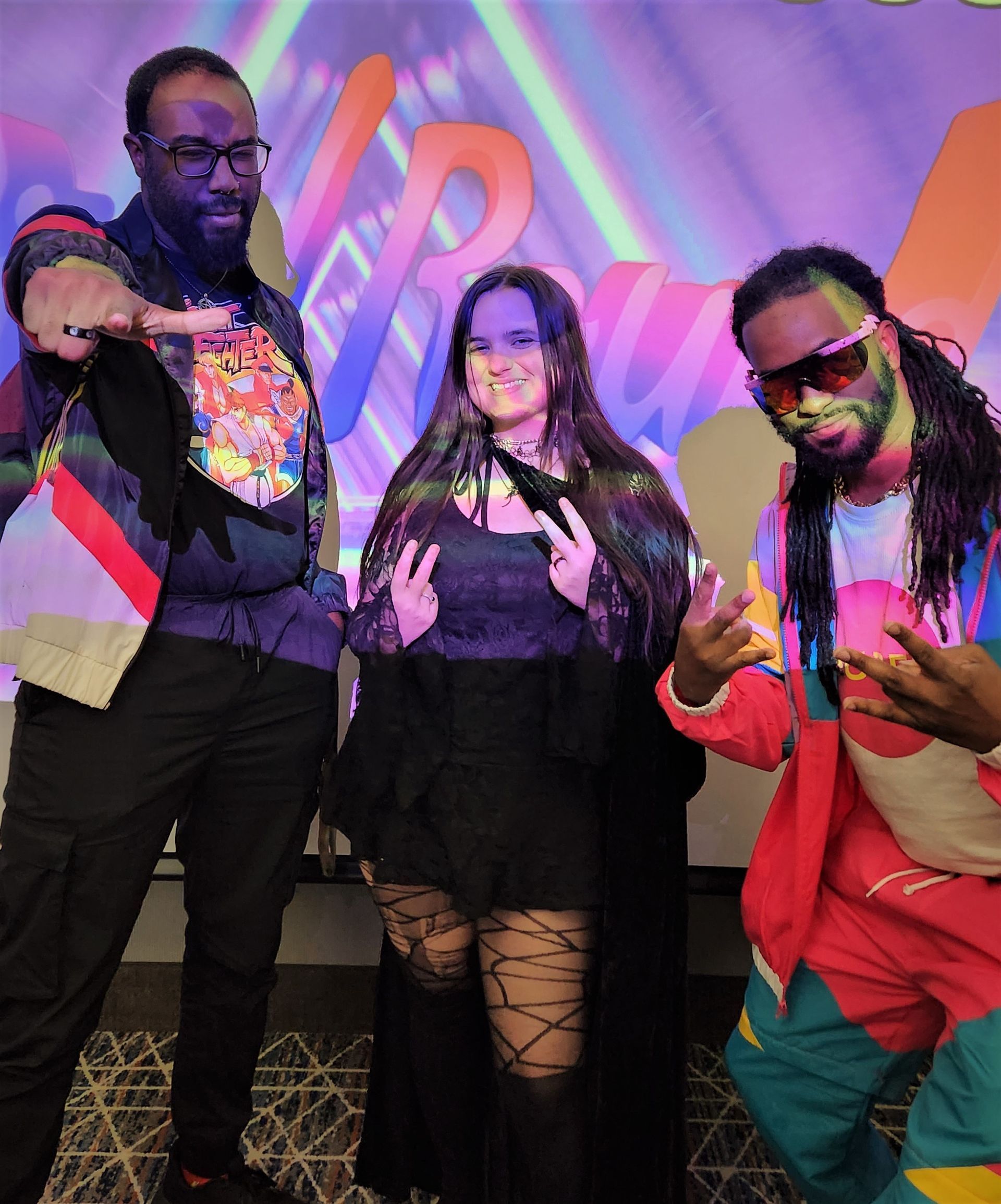 Three people pose in front of a colorful backdrop. Man in glasses points, woman makes peace signs, man in shades gestures.