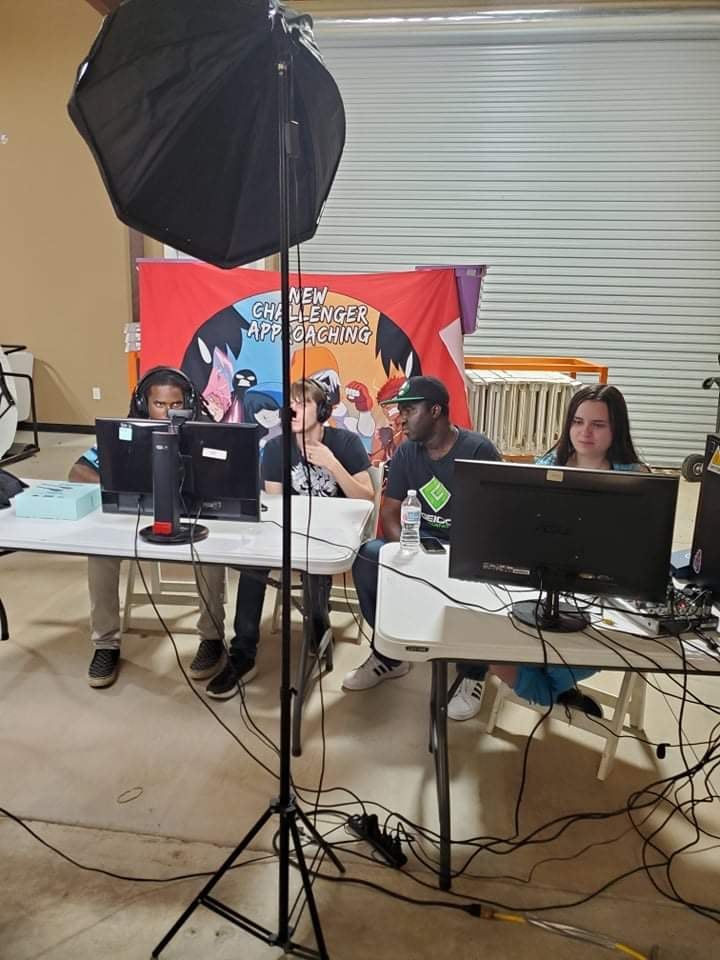 Four people sit at tables with computers, in front of a banner and studio light.